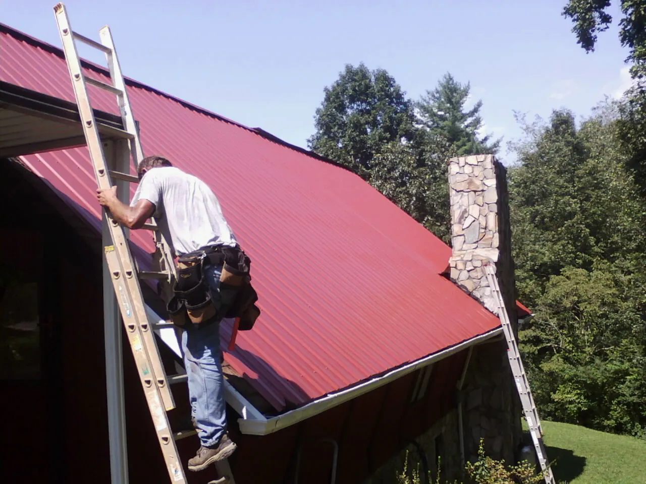 Roofer on a ladder beside a red metal roof and stone chimney — Soffit & Fascia Repair by Roofing Brothers in Dahlonega